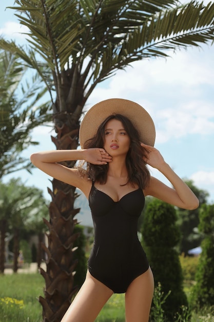 Wunderschöne junge Frau mit gebräuntem sexy Körper im schwarzen Bikini und im Strohhut, die Sommerzeit nahe Palmen genießen