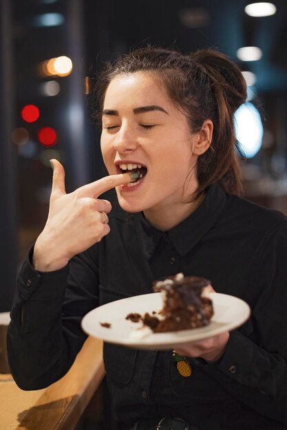 Wunderschöne lächelnde junge Frau mit dunklem Haar, die Kuchen isst und Kaffee in einer Cafeteria trinkt, die abends am Fenster sitzt, selektiver Fokusgeräuscheffekt