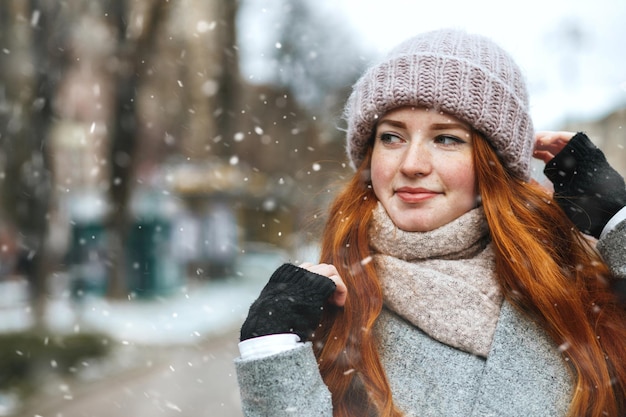 Wunderschöne rothaarige Frau trägt einen Mantel, der während des Schneefalls in der Stadt spazieren geht. Platz für Text