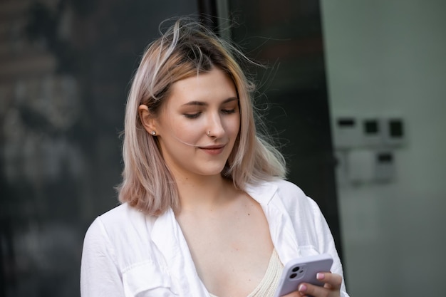 Wunderschöne schöne junge Frau mit blonden Haaren, die auf dem Smartphone auf dem Hintergrund der Stadtstraße Nachrichten sendet. Hübsches Mädchen, das ein Smartphone-Gespräch auf der Stadtstraße führt