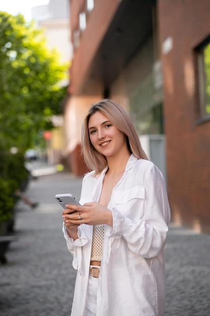 Wunderschöne schöne junge Frau mit blonden Haaren, die auf dem Smartphone auf dem Hintergrund der Stadtstraße Nachrichten sendet. Hübsches Mädchen, das ein Smartphone-Gespräch auf der Stadtstraße führt