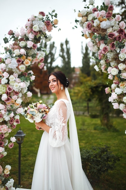 Wunderschöne Verlobte im weißen Hochzeitskleid mit Blumenstrauß in der Nähe des Hochzeitsbogens aus Blumen