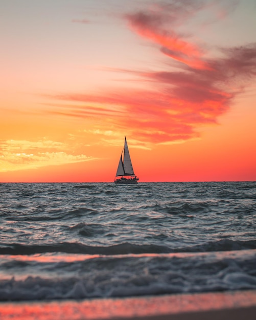 Wunderschöne vertikale Aufnahme einer Yacht, die bei orangefarbenem Sonnenuntergang im offenen Meer segelt