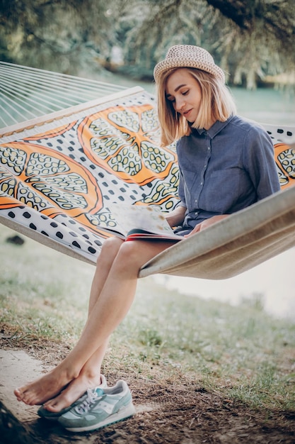 Wunderschönes blondes Hipster-Mädchen in blauem Hemd und Vintage-Hut, das ein Buch auf einer Hängematte liest. Junge Frau auf einem Campingausflug im Waldsee im Hintergrund