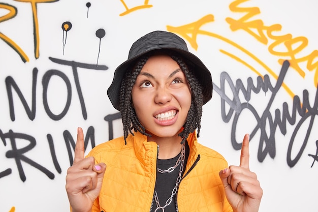 Wunderschönes ethnisches Hipster-Mädchen mit frechen Gesichtsausdrücken zeigt etwas gegen die Graffiti-Wand in modischem Outfit