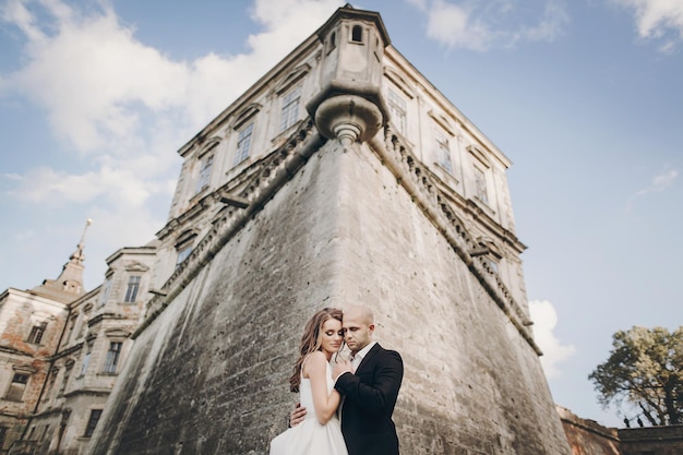 Foto wunderschönes hochzeitspaar, das sich im sonnenlicht in der nähe des alten schlosses in einem wunderschönen park umarmt. stilvolle schöne braut und bräutigam, die sich sanft auf dem hintergrund des alten gebäudes und der natur umarmen