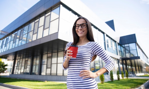 Wunderschönes intelligentes Mädchen mit Brille steht vor dem modernen Gebäude und hält eine rote Kaffeetasse in der rechten Hand, mit der linken Hand auf einer Hüfte