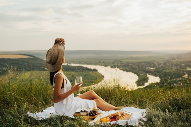 Wunderschönes junges brünettes Mädchen in einem weißen Sommerkleid und Strohhut, die ein Picknick an einem malerischen Ort genießen.