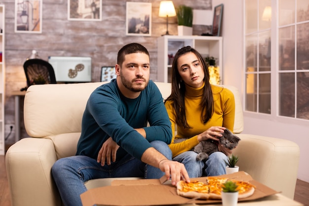 Wunderschönes junges Paar, das Pizza isst, während es im Wohnzimmer auf dem Sofa sitzt?