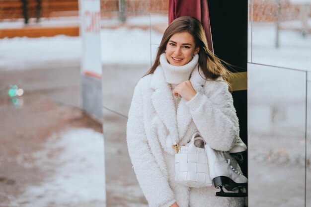 Wunderschönes weibliches Model im weißen Mantel und weißen Pullover hält den Schlittschuh zufrieden, nachdem er auf der Eisbahn gelaufen ist