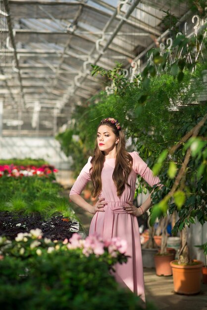 Wunderschönes weibliches Modell in einem langen rosafarbenen Kleid posiert zwischen den vielen Blumen im Gewächshaus