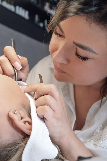 Wundervoller Wimpernmacher sitzt mit einer Pinzette über der Kundin und klebt sorgfältig künstliche Wimpern. Konzentrierte Brünette blickt der Kundin ins Gesicht und führt eine Wimpernverlängerungsprozedur durch