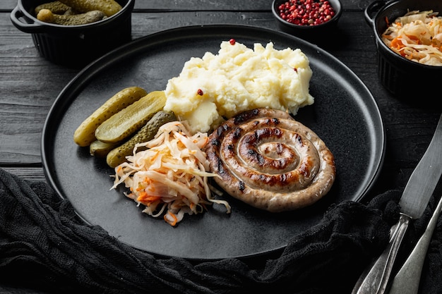 Wurst oder Bratwurst mit fermentiertem Kohl, eingelegten Gurken und Gewürzen auf schwarzem Holztischhintergrund