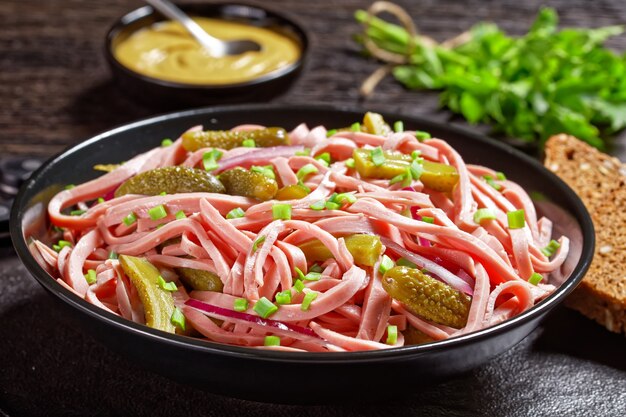 Wurstsalat alemán de salchicha lyoner alemana cocida con pepinos encurtidos, rodajas de cebolla roja, aderezo de vinagre servido en un cuenco negro sobre un fondo de madera oscura con mostaza, vista superior, primer plano