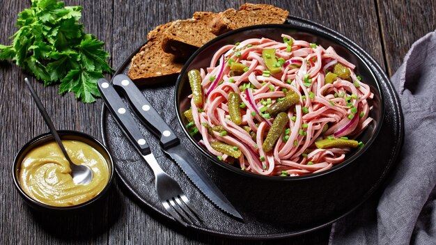 Wurstsalat alemán de salchicha lyoner alemana cocida con pepinos encurtidos, rodajas de cebolla roja, aderezo de vinagre servido en un cuenco negro sobre un fondo de madera oscura con mostaza, vista superior, primer plano