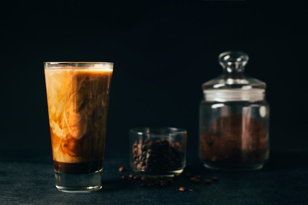 Foto xaclose de um copo de café com leite contra o fundo de um copo de grãos de café e uma jarra de café moído em um fundo preto