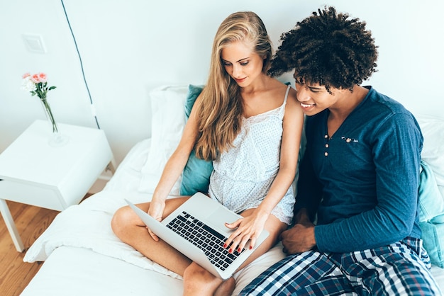 XAYoung schönes gemischtrassiges Paar entspannt im Bett liegend auf dem Computer