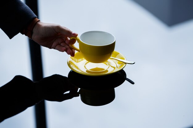Xícara amarela para café ou chá com um pires nas mãos de uma garota no fundo de uma mesa de vidro.