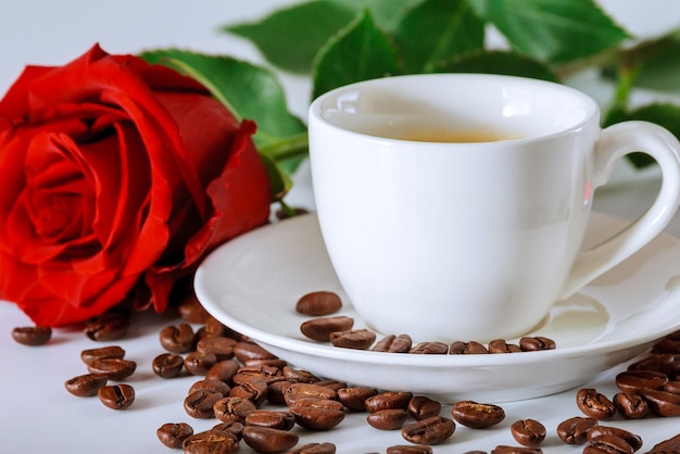 Xícara branca de café preto com uma rosa vermelha e grãos de café espalhados em uma mesa branca.