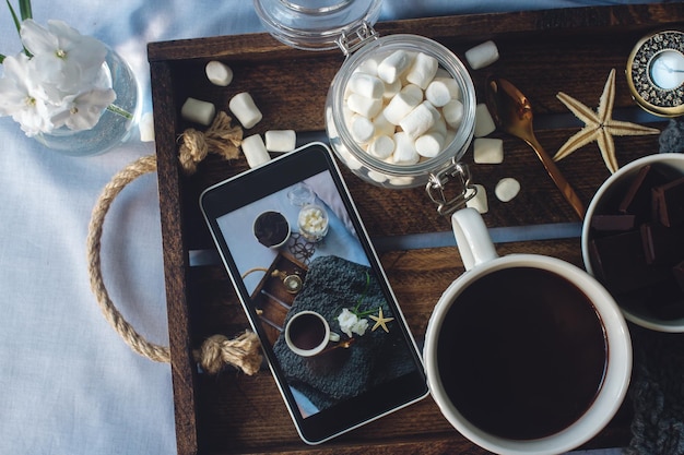 Xícara de cacau caseiro com flores de chocolate marshmallow e smartphone na bandeja de madeira rústica na cama aconchegante fim de semana manhã blogueiro estilo de vida vista superior foco seletivo