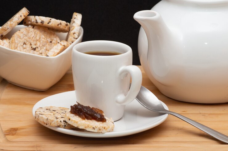 Foto xícara de café ao lado de biscoitos de arroz vegan, uma tigela cheia de biscoitos e uma cafeteira sob a mesa de madeira