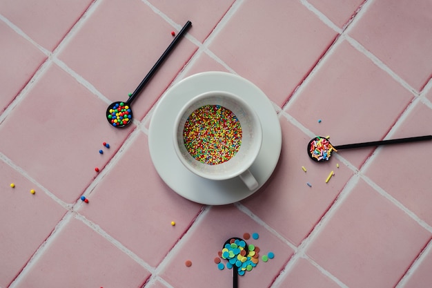 Xícara de café branca cheia de granulado de doce e uma colher preta em um fundo de balcão de ladrilhos rosa