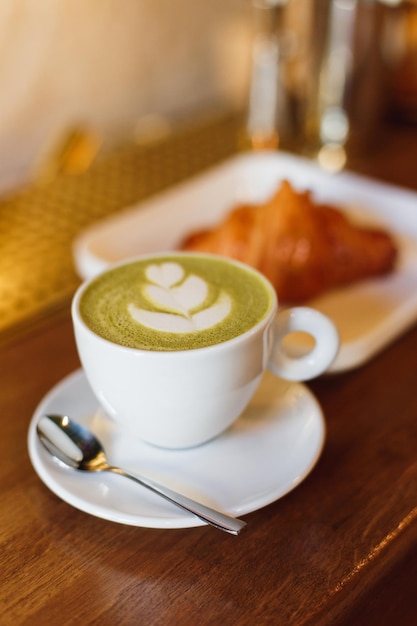 Xícara de café branca com matcha cappucsino e um prato com um croissant em um balcão de bar de madeira