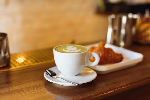 Xícara de café branca com matcha cappucsino e um prato com um croissant em um balcão de bar de madeira