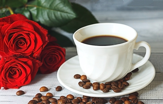 Xícara de café branca em grãos de café e rosas vermelhas no conceito de café da manhã romântico de fundo cinza claro