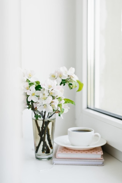 Xícara de café branca em um prato branco uma pilha de livros e um vaso de flores no parapeito da janela Aconchegante Páscoa primavera natureza morta