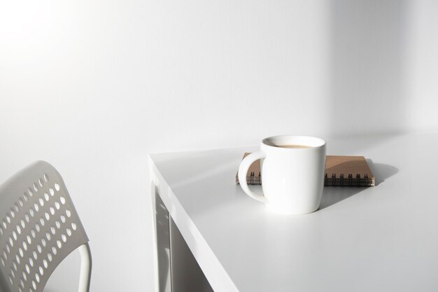 Xícara de café branca, um caderno sobre uma mesa branca, há uma cadeira ao lado