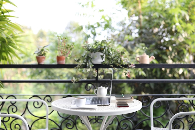 Xícara de café branco e laptop e com vaso de plantas na mesa de metal branco na varanda