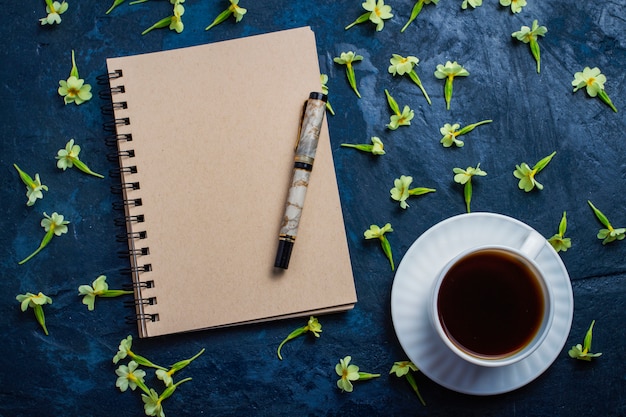 Xícara de café, caderno e flores sobre um fundo azul escuro. Vista plana e superior