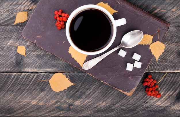 Foto xícara de café com colher e açúcar, livro antigo, folhas de outono e sorveira seca em fundo de madeira