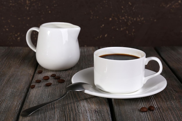 Xícara de café com creme na jarra de leite e grãos de café na mesa de madeira em fundo escuro