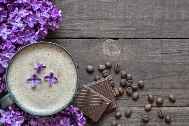 Xícara de café com grãos de café, flores de chocolate e lilás sobre a mesa de madeira rústica