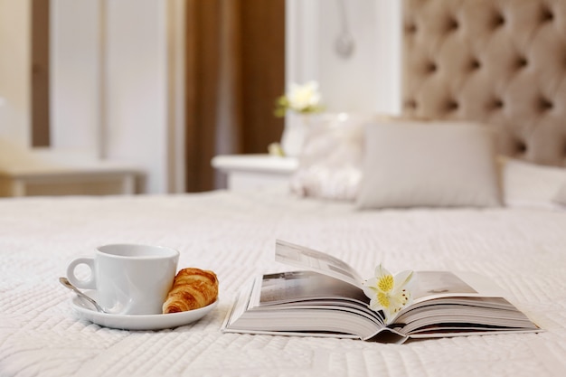 Xícara de café, croissant e um livro na cama em um quarto confortável e luminoso