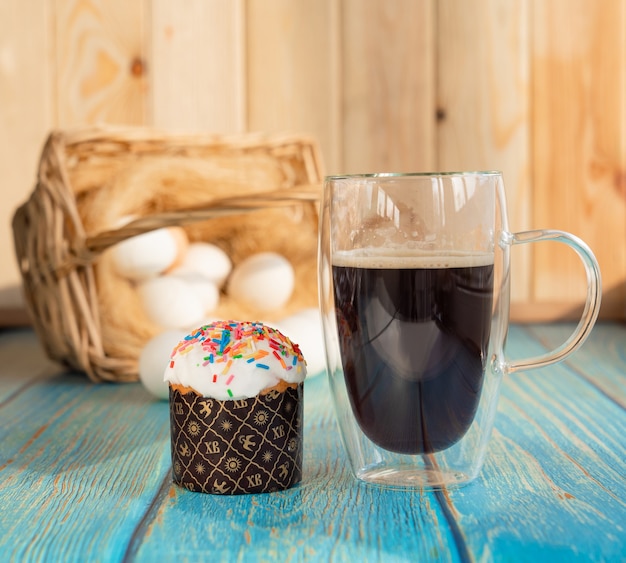 Xícara de café de vidro. Kulich de Páscoa e ovos em uma cesta no feno. mesa de madeira turquesa.