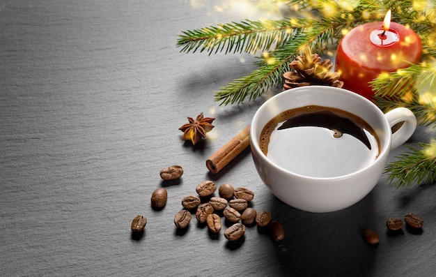 Xícara de café e grãos de café com ramo de abeto de decoração de natal e vela vermelha acesa na mesa de pedra escura texturizada.