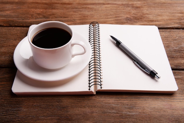 Xícara de café em pires com caderno e caneta no fundo da mesa de madeira