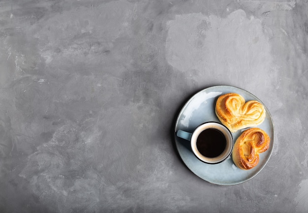 Xícara de café expresso com dois pãezinhos doces em forma de coração no prato sobre concreto cinza. Natureza morta minimalista. Vista do topo.