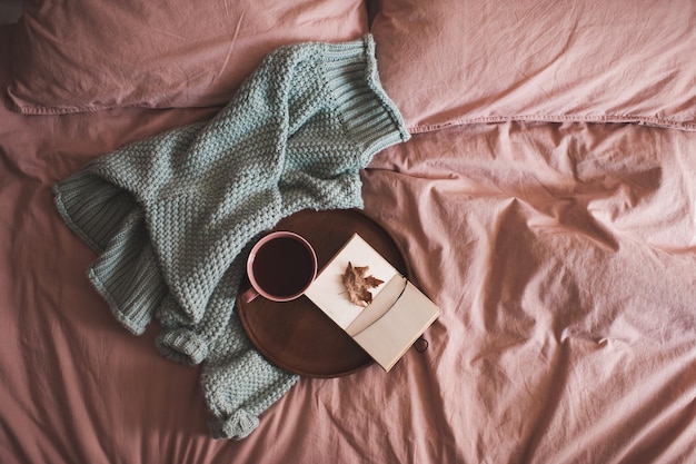 Xícara de café fresco com o caderno de desenho aberto com folhas caídas de outono na bandeja de madeira e a camisola de malha na cama close-up. Bom Dia.