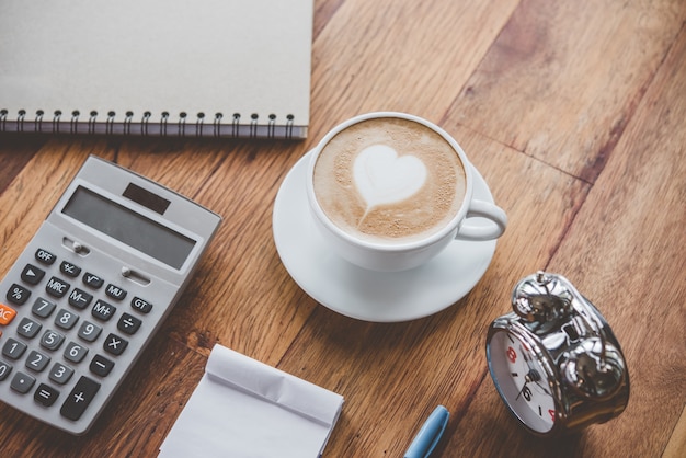 xícara de café latte art forma de coração com ferramentas de escritório na mesa
