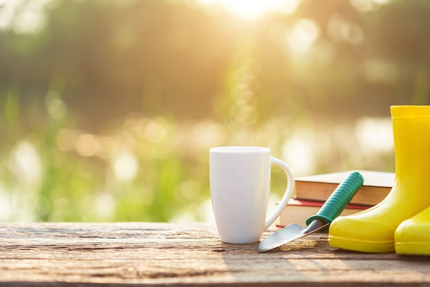 Xícara de café, livro e equipamento de jardim na mesa de madeira com luz solar no tempo da manhã