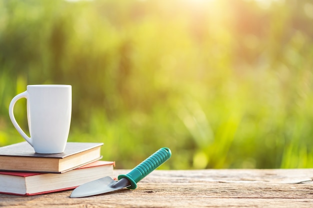 Xícara de café, livro e equipamento de jardim na mesa de madeira com luz solar no tempo da manhã