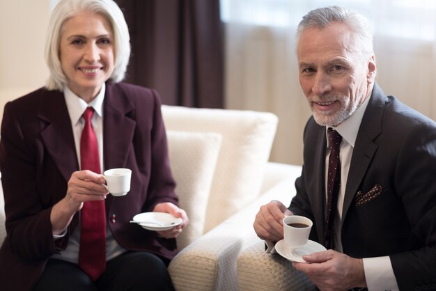 Xícara de café. Mulher de negócios alegre e encantada sorrindo e sentada no escritório enquanto bebe um café com seu colega e expressa felicidade