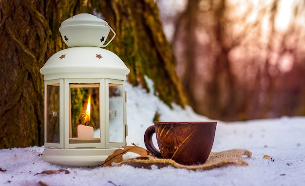 Xícara de café na floresta de inverno ao lado da lanterna com vela perto de uma árvore velha à noite