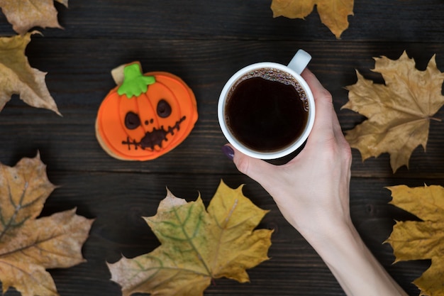 Xícara de café na mão feminina e abóbora de biscoitos de gengibre para o feriado de Halloween Folhas de outono Vista superior