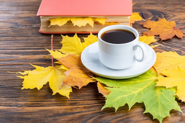 Xícara de café na mesa de madeira com livro e folhas de bordo outonais coloridas