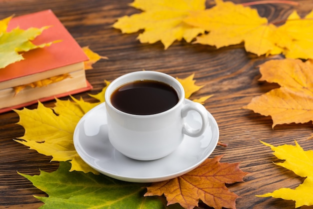 Xícara de café na mesa de madeira com livro e folhas de bordo outonais coloridas